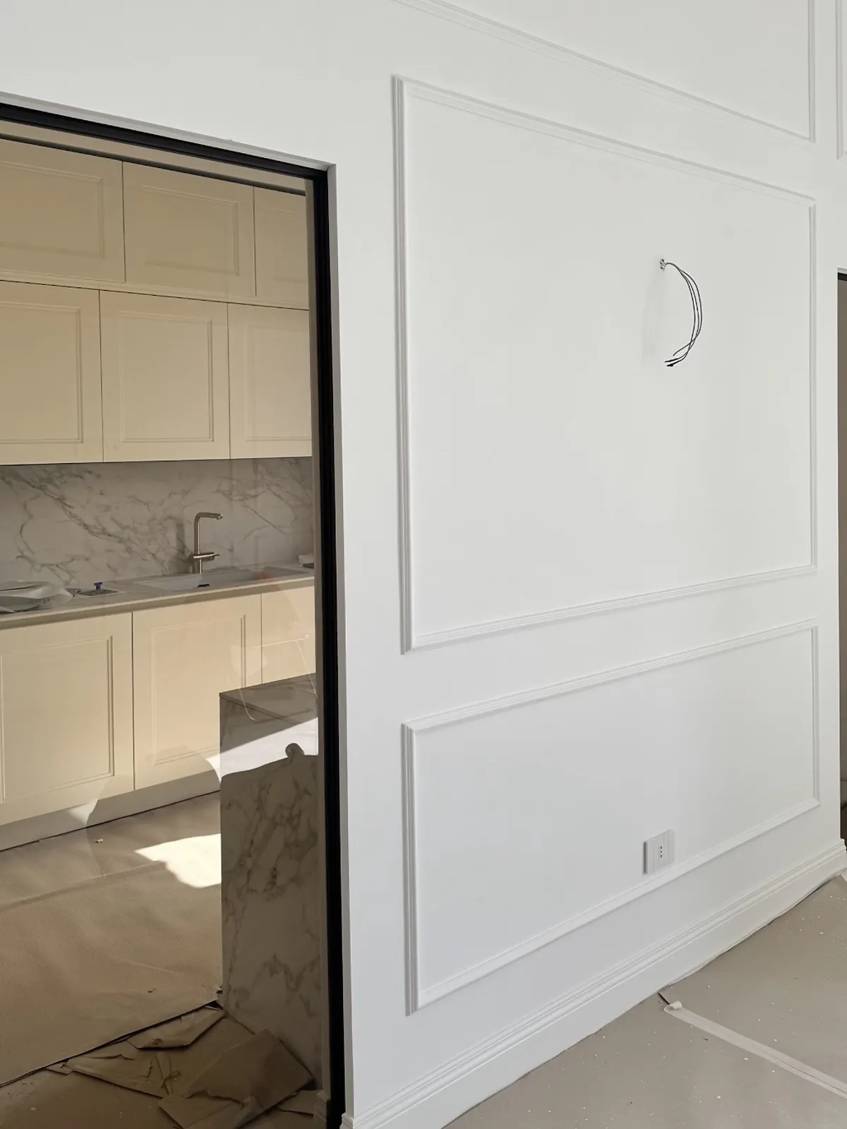 La stessa parete finita: boiserie bianca, porta in vetro con profilo nero e cucina color crema con marmo